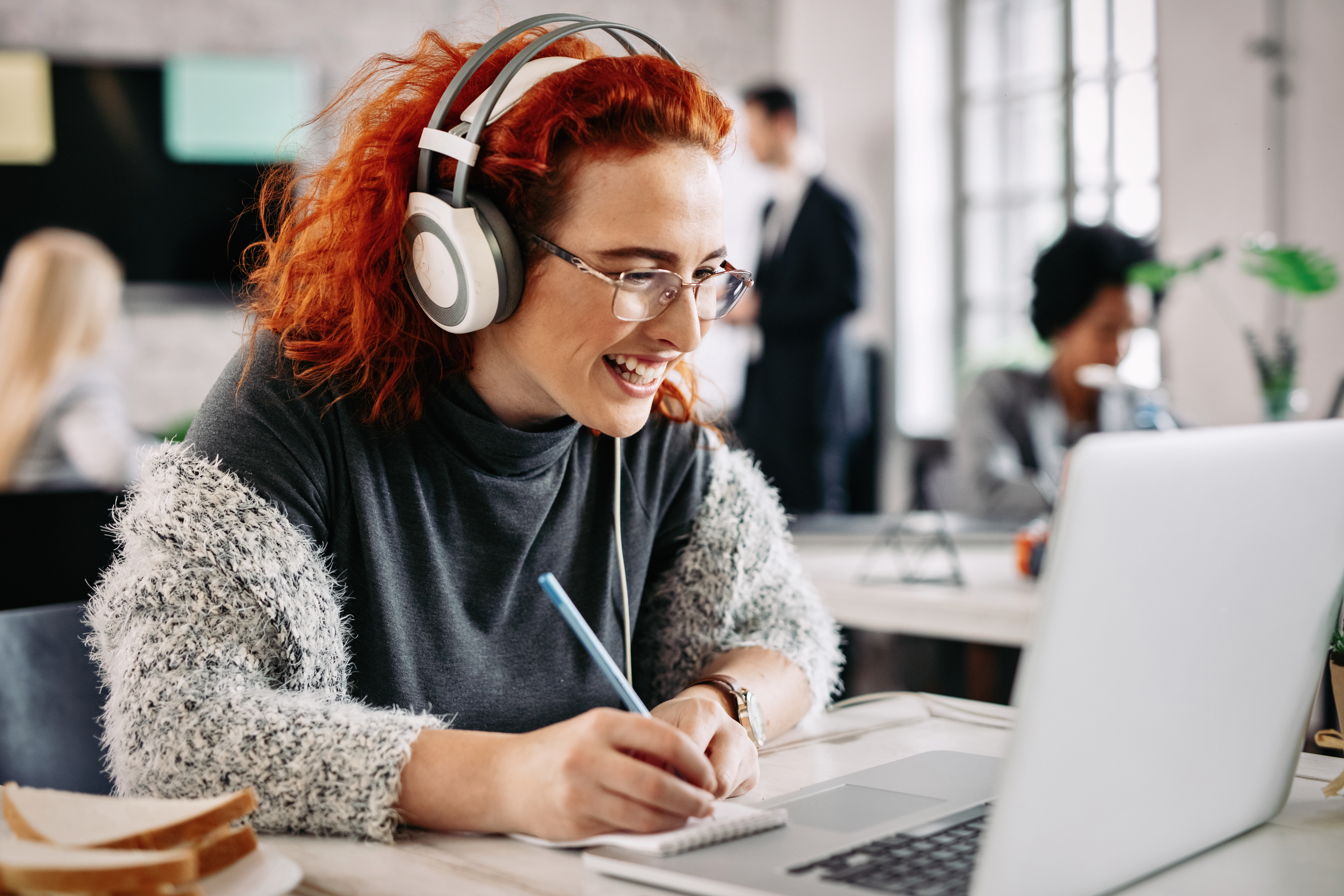 cheerful-female-entrepreneur-working-computer-taking-notes-her-notepad-while-listening-music-headphones-office-there-are-people-background
